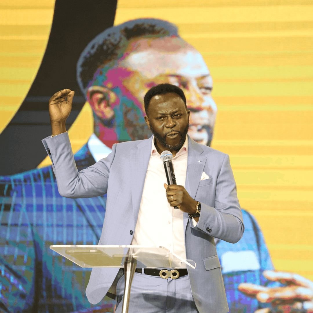 A minister in a light grey suit preaching in front of a bright yellow screen